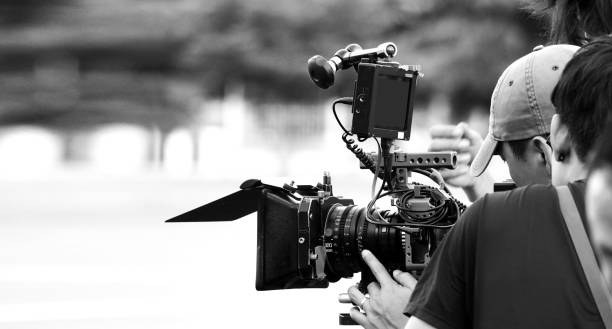 istock Back angle view of high definition digital video camera which recording or shooting tv commercial production on pan tilt shift professional head tripod at outdoor location and film crew team with set. 2107262406