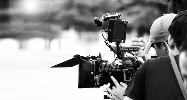 istock Back angle view of high definition digital video camera which recording or shooting tv commercial production on pan tilt shift professional head tripod at outdoor location and film crew team with set. 2107263488