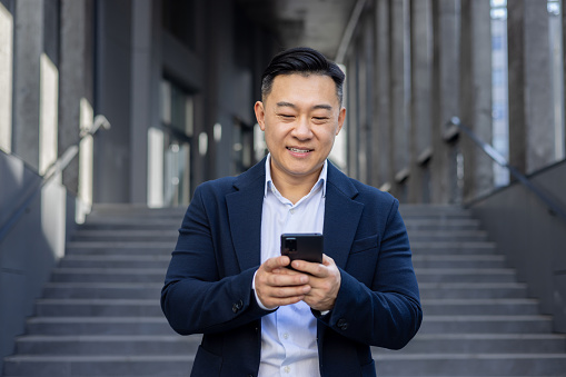 Smiling Asian executive using mobile phone for texting while standing on stairs outside a modern building.