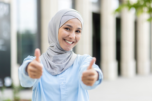 A cheerful, young woman wearing a hijab gives a double thumbs up with a broad, confident smile, signaling approval and success.
