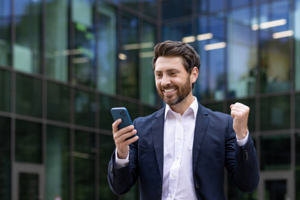 istock Enthusiastic businessman celebrating success with smartphone outside modern office 2107394800
