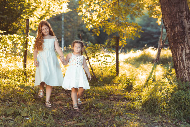 Two little girls, sisters walking outdoors in white dresses, holding hands. Copy space