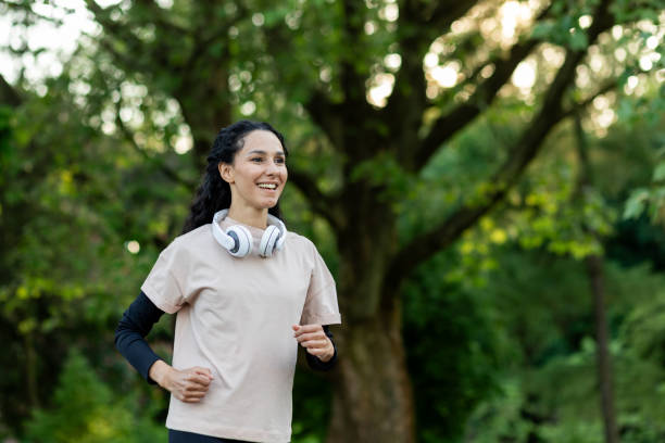 istock Active female in tshirt over long sleeve with headset over neck exercising outdoors during daytime in green area. Positive lady doing jogging in public park for keeping fit and healthy body 2107412856