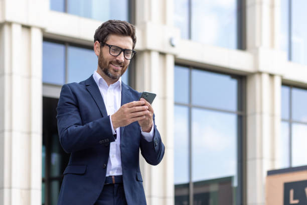 istock Smiling businessman in suit using smartphone outdoors with office building 2107462711