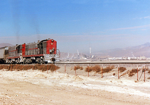 Freight train departing Trona, California, with several spewing smoke stacks within industrial  complex in background.