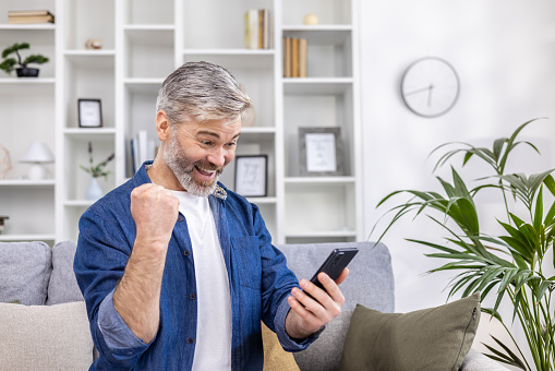 Mature gray haired man alone at home using phone and celebrating victory success happy holding hand up, adult person sitting on sofa in living room on sunny day.