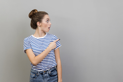 Wow, look at ads here. Portrait of astonished woman in striped T-shirt looking away with surprised amazed expression, pointing aside at copy space. Indoor studio shot isolated on gray background.