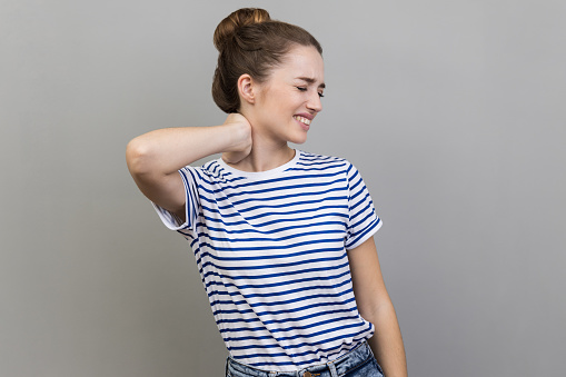 Portrait of beautiful upset unhealthy sick woman wearing striped T-shirt standing and holding her painful neck, feels bad, frowning face. Indoor studio shot isolated on gray background.