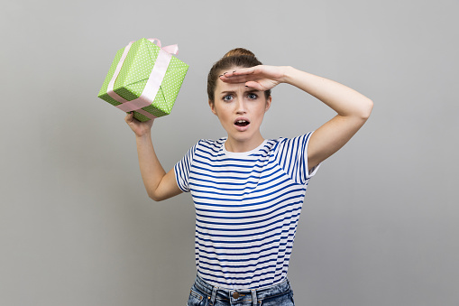 Portrait of shocked surprised woman wearing striped T-shirt holding far away with open mouth and hand over forehead, holding present box. Indoor studio shot isolated on gray background.