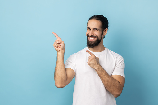 Portrait of smiling man with beard in white T-shirt pointing finger aside, showing copy space for commercial text, blank wall with idea presentation. Indoor studio shot isolated on blue background.