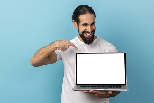 Portrait of man with beard wearing white T-shirt standing holding laptop with blank screen and smiling at camera, internet advertising. Indoor studio shot isolated on blue background.