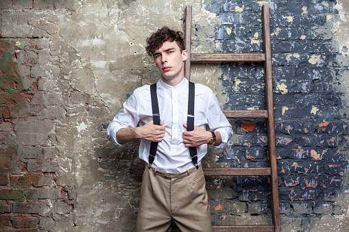 Portrait of serious confident man in white shirt and beige pants holding suspenders, leaning on wooden ladder, looking at camera, posing on brick wall background. Indoor studio shot.