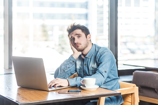 Portrait of sad depressed young man freelancer in blue jeans shirt working on laptop, feels bored, leaning on his hand, looking at camera. Indoor shot near big window, cafe background.
