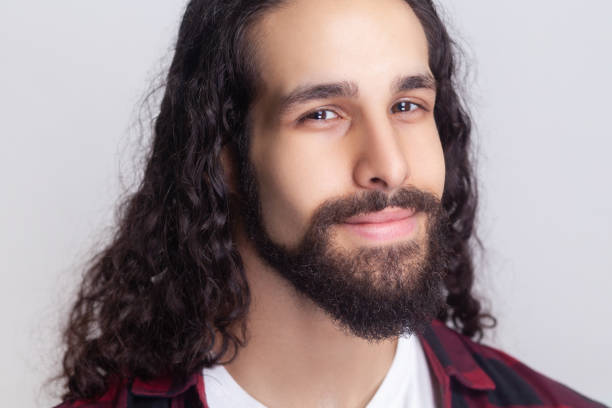 Closeup portrait of calm handsome bearded man with long curly hair in checkered red shirt looking at camera with smile, expressing positive emotions. Indoor studio shot isolated on gray background.