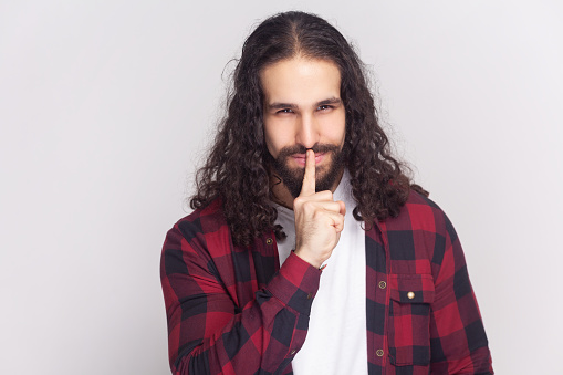 Mysterious bearded man with curly hair in checkered red shirt makes shush gesture, shows hush sign, asks not tell secret, presses index finger to lips. Indoor studio shot isolated on gray background.