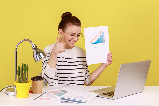Delighted woman holding growth diagram, showing thumb up to laptop screen, satisfied with financial and economic growth of his business. Indoor studio studio shot isolated on yellow background.