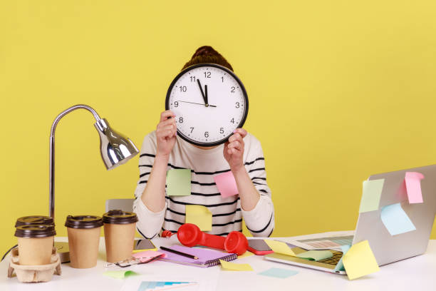 Woman office worker covered with sticky notes sitting at workplace hiding face behind big wall clock