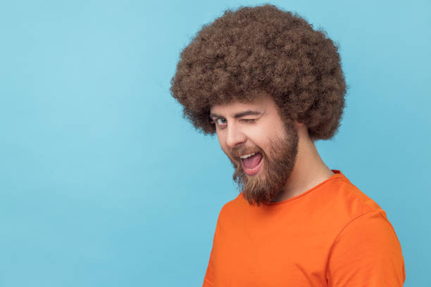 Man in good mood, smiling broadly and winking at camera with toothy smile. Portrait of flirting man with Afro hairstyle wearing orange T-shirt in good mood, smiling broadly and winking at camera with toothy smile. Indoor studio shot isolated on blue background. men hairstyle afro cool stock pictures, royalty-free photos & images