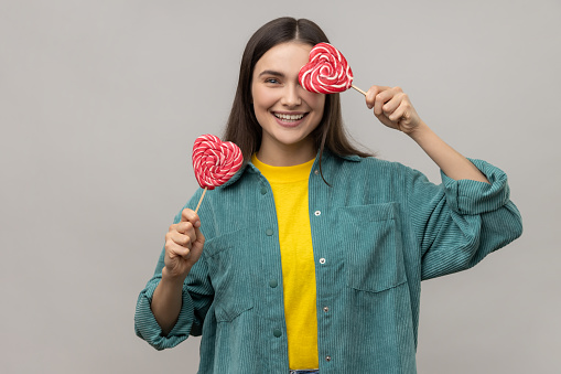 Childish woman with dark hair covering eye with heart shape lollipops, hiding, having fun with sweets, wearing casual style jacket. Indoor studio shot isolated on gray background.