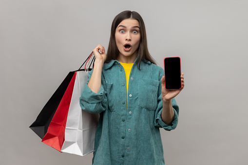 Amazed woman standing with astonished look showing shopping bags and smart phone with blank screen for advertisement, wearing casual style jacket. Indoor studio shot isolated on gray background.