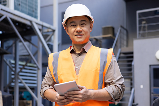 A professional industrial worker wearing a high-visibility vest holds a tablet while standing at an industrial site.
