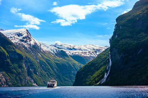 Cruising the majestic Geirangerfjord in the gorge inlet between steep snowcapped mountains lined with lush green trees, foliage and powerful waterfalls and flowing water from melted snow on a mostly blue sky with partial clouds