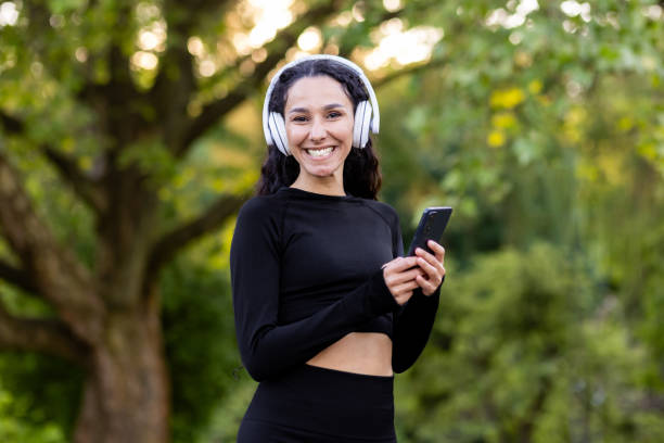 istock Active brunette woman wearing wireless headset and black sportswear while looking at camera with smartphone in hand. Smiling female doing daily exercises to music on background of public park 2107764937