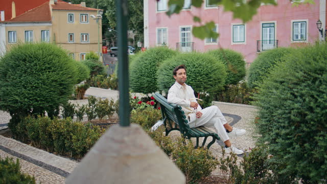 Romantic man waiting girlfriend at green park bench. Boyfriend holding flower