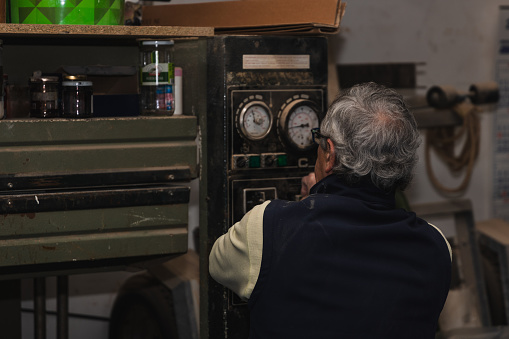 elderly carpenter, seen from behind, as he observes the gauges of an old wood press machine to monitor time and pressure in his workshop. The scene embodies a blend of experience and precision, highlighting the timeless craftsmanship and attention to detail in woodworking