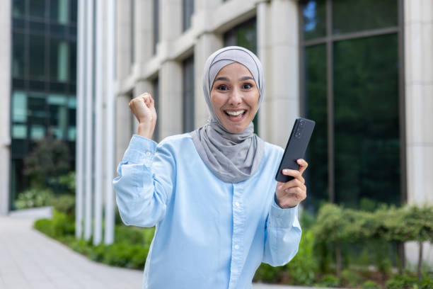 istock Excited muslim female cheering with mobile phone in raised hands while walking down business district. Emotional woman receiving unexpected pleasant notification about salary transfer in banking app 2107798431