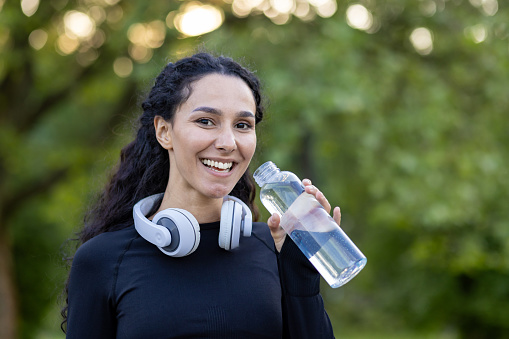 Joyful athletic woman resting and drinking water in park with headphones around her neck, smiling at the camera.