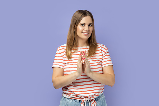 Cunning blond woman wearing striped T-shirt gesticulating and thinking over devious sly plan of revenge, scheming and conspiring villain plan. Indoor studio shot isolated on purple background.
