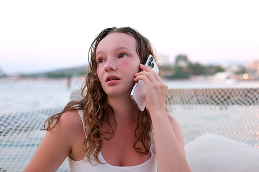 A woman talks on the phone, ordering delivery, communicates with friends, uses social networks and the Internet. Relaxing on beach emotions of young girl burnt skin red cheeks wet hair.