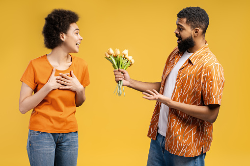 Attractive African American couple, bearded young man giving yellow tulips to excited woman with curly hair, standing isolated on yellow background. Concept of celebration, birthday