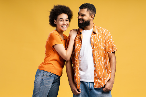 Smiling African American couple, man and woman wearing stylish casual clothes, hugging and looking at camera. Attractive boyfriend and girlfriend standing isolated on yellow background. Summer concept