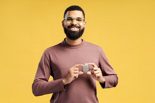 Smiling, attractive African American man holding pacemaker looking at camera. Patient showing cardioverter defibrillator standing isolated on yellow background. Concept of health care, support