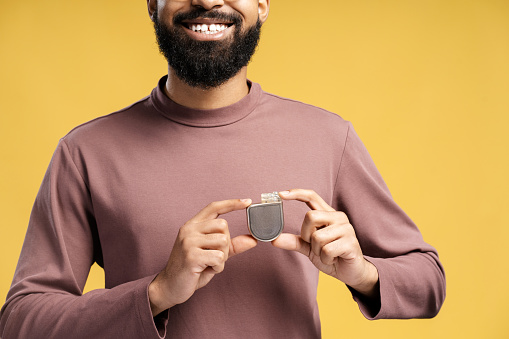 Happy handsome African American man holding pacemaker looking at camera closeup. Patient showing cardioverter defibrillator standing isolated on yellow background. Concept of health care, support