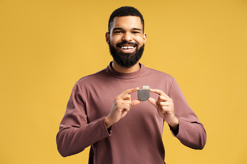 Smiling African American man holding pacemaker, cardioverter defibrillator looking at camera. Attractive patient standing isolated on yellow background. Health care concept, cardio treatment