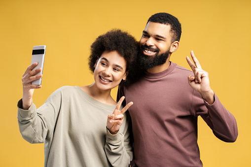 Attractive smiling African American man and woman, boyfriend and girlfriend using mobile phone taking selfie, showing victory sign, standing isolated on yellow background. Online technology concept