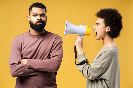 Unhappy couple, African American woman holding loudspeaker quarreling, shouting standing isolated on yellow background. Concept of abusive relationships