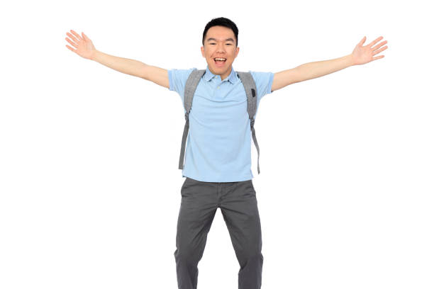 A young adult Asian high school or university student is posing with stretched arms happily. He is wearing a light blue polo shirt and wearing a grey backpack. The background is white and isolated in a studio.