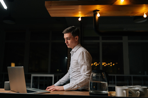 Tired young businessman working laptop in evening office. Focused business man reading computer standing by desk. Overworked entrepreneur looking screen solving tasks in creative night space.