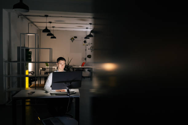 istock Remote view of focused young office worker male working at workplace on computer at late night in dark room. Successful entrepreneur checking finances on internet 2107893770