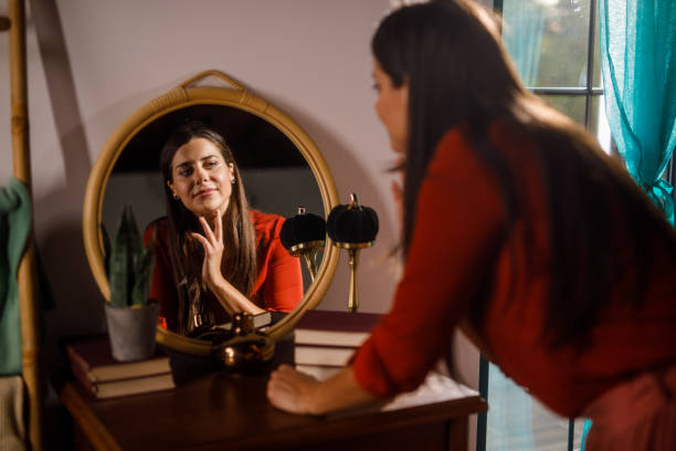 Selective focus shot of smiling young woman looking in the mirror and fixing her hair and make up when getting ready.