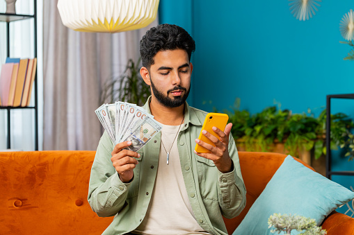 Planning budget. Rich happy Indian man use smartphone calculator app counting money cash, calculate domestic bills at home. Guy satisfied of income earnings win saves money for planned vacation gifts