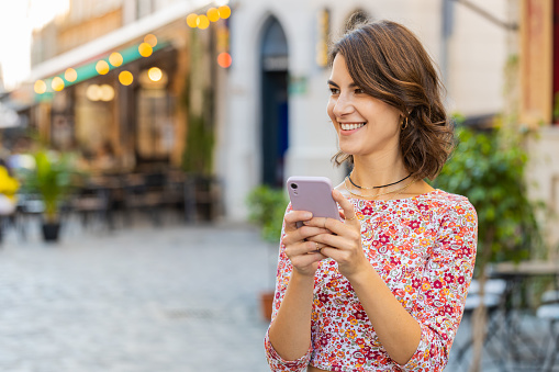 Smiling pretty young woman using smartphone typing text answering messages chatting online looking mobile screen social media app. Girl tourist walking in city street outdoors holding phone in hands