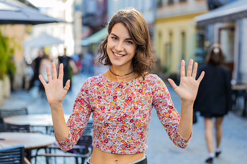 Hello. Young woman smiling friendly at camera, waving hands gesturing hi, greeting or goodbye, welcoming with hospitable expression outdoors. Happy lovely adult girl standing in urban city town street