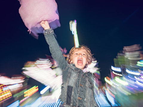 Cheerful boy with sweet cotton candy shouting and looking away while standing near illuminated attraction ride in amusement park in evening time