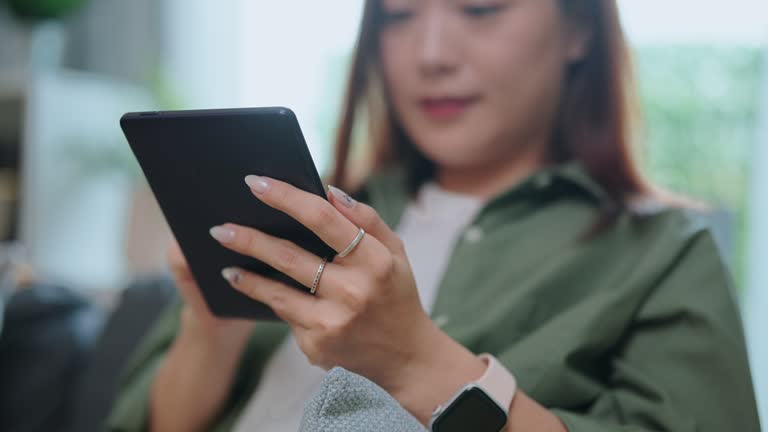 Asian woman relaxing reading on E-book holding a tablet displaying text on sofa in living room at home ,modern lifestyle