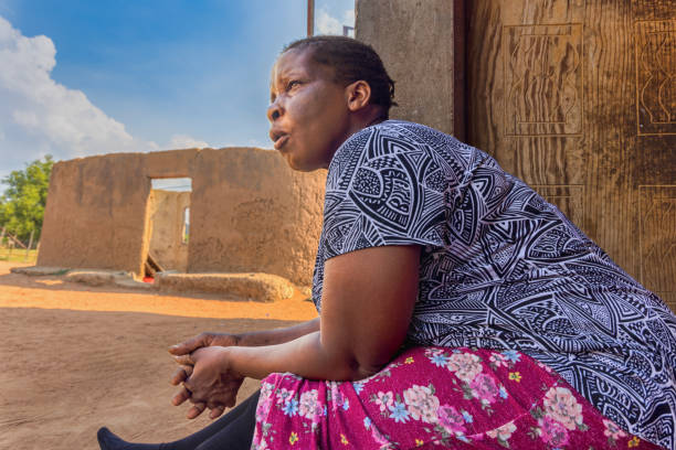 village african woman with braids in front of her home, sited on the veranda, ruins of mud house in the background
